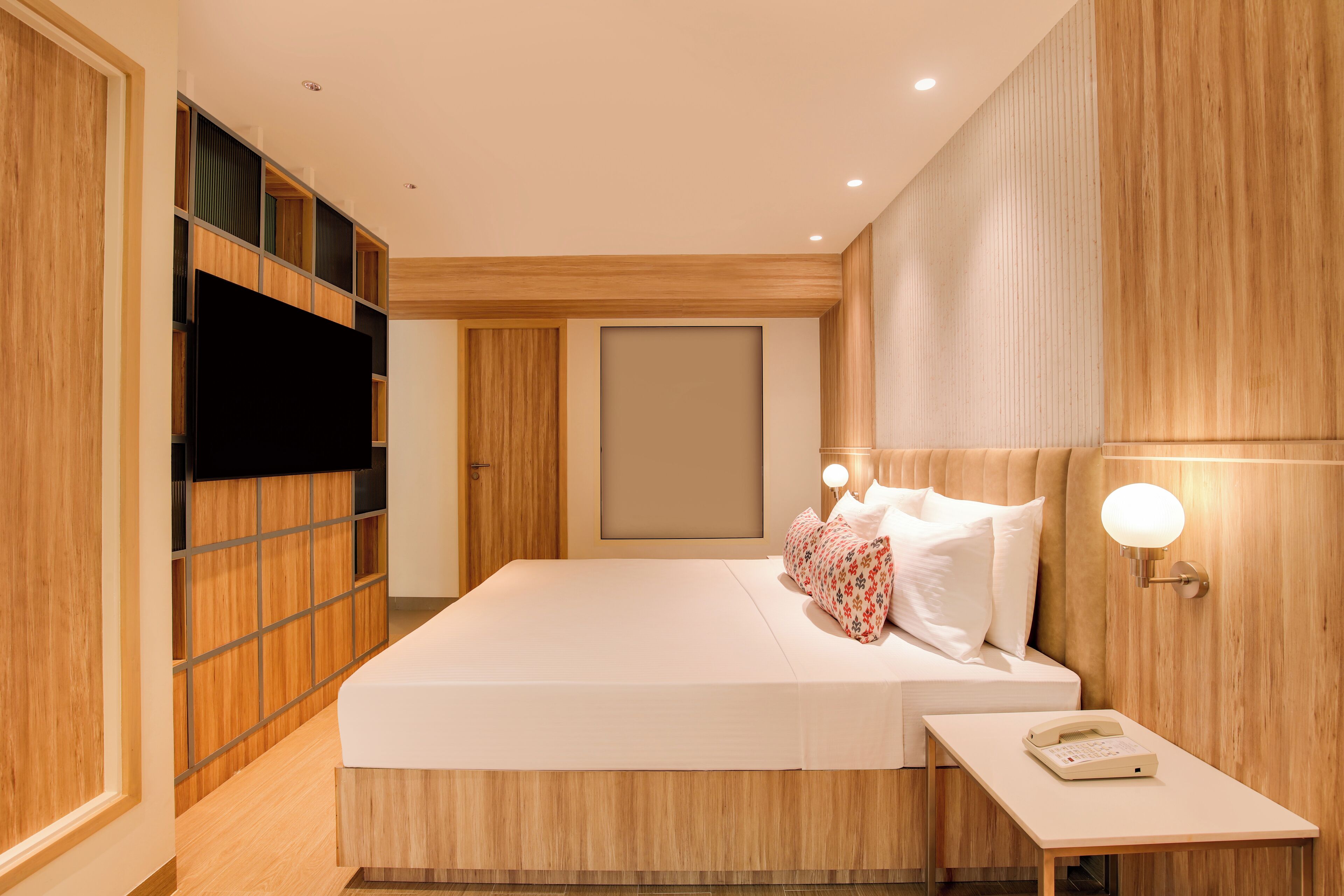 A modern hotel room with a large bed, wooden paneling, a flat-screen TV, and a bedside table with a lamp.