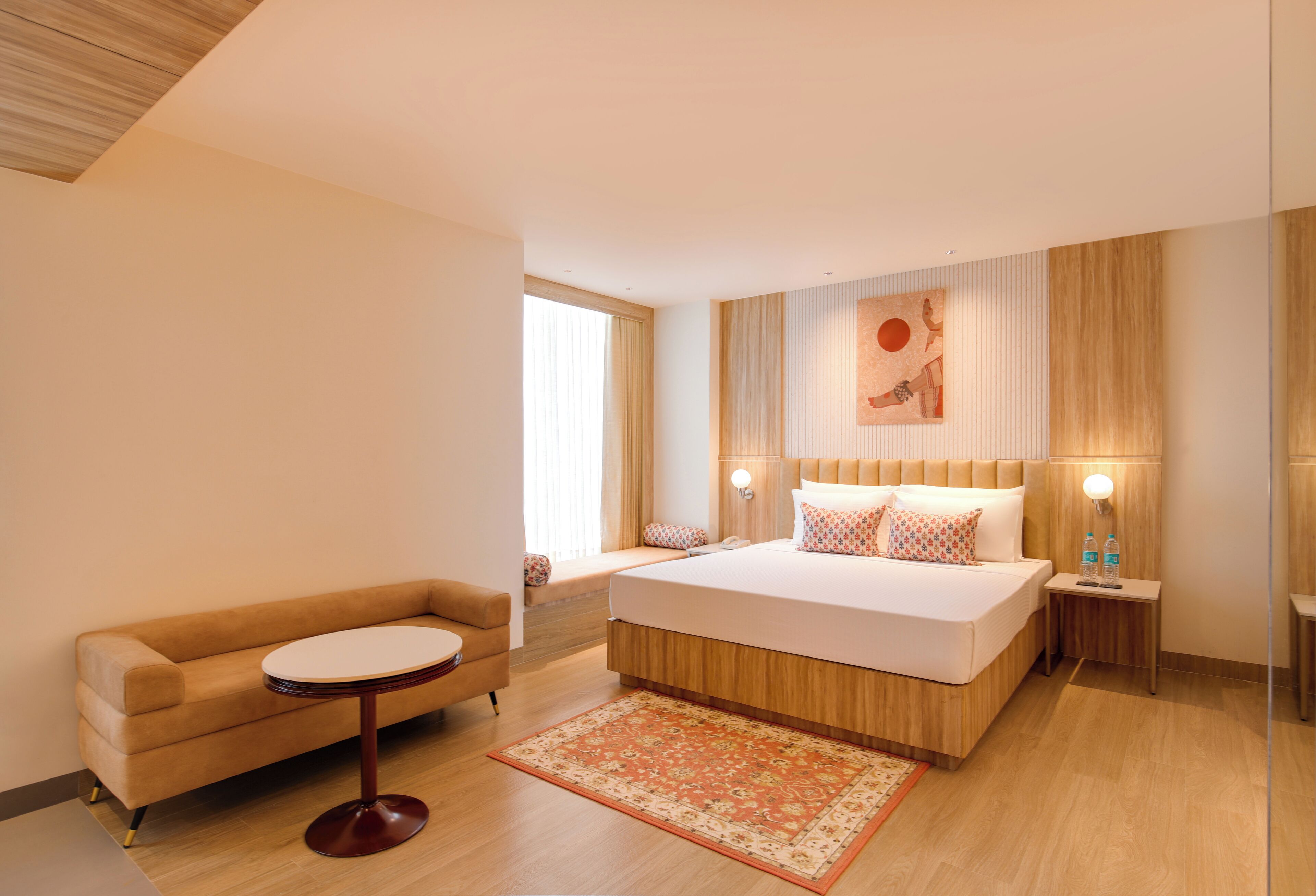Une chambre d’hôtel moderne avec un grand lit, un canapé, une petite table ronde et une table de chevet sur laquelle sont posées deux bouteilles.