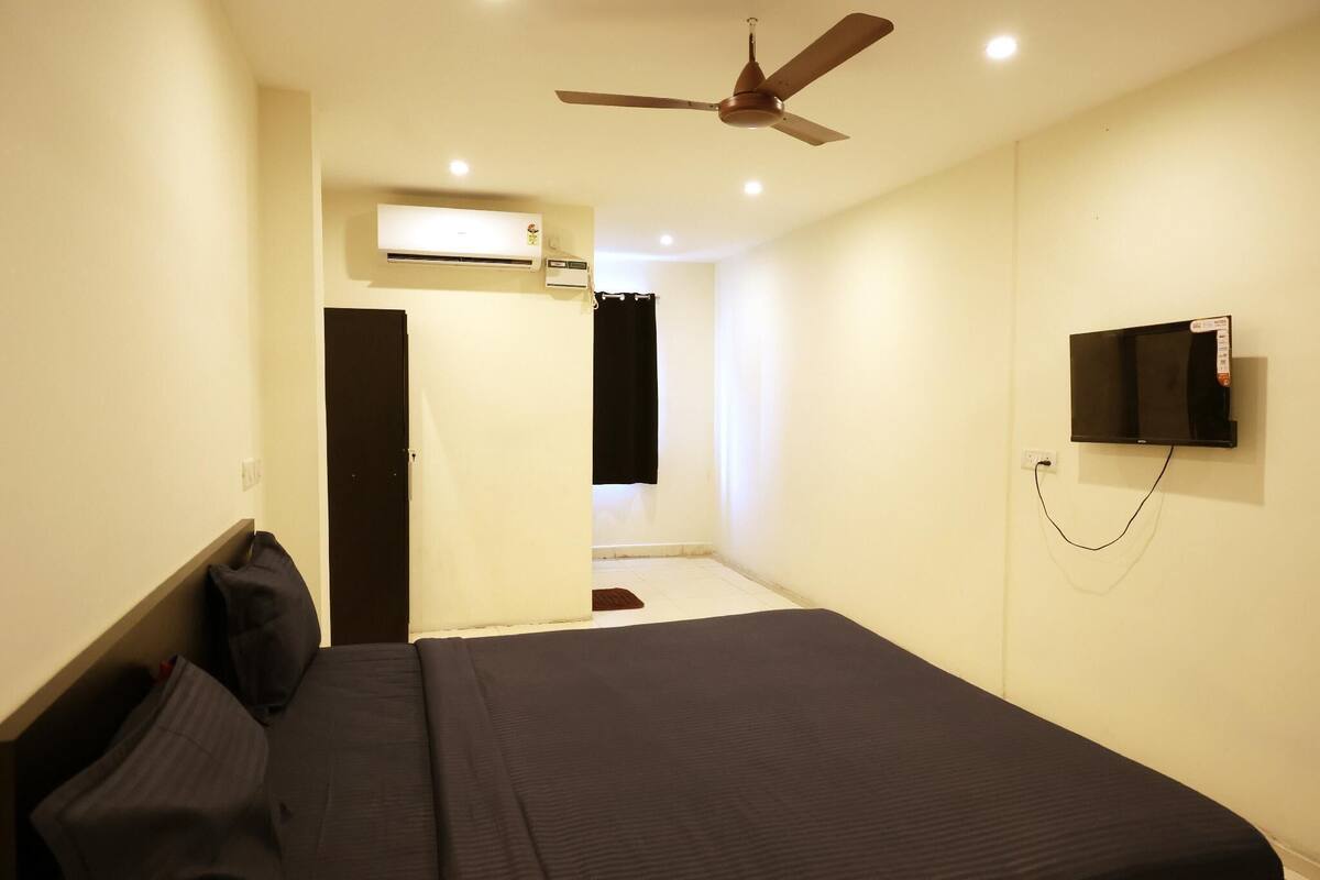 Una habitación de hotel con una cama, ventilador de techo, unidad de aire acondicionado, televisión montada en la pared y una puerta que da a un baño.