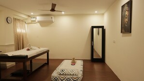 Quarto com mesa de massagem, cama pequena com colchão estampado, ventilador de teto, ar-condicionado, relógio e uma obra de arte emoldurada na parede.