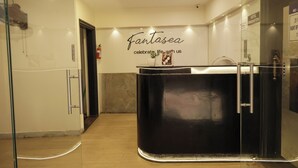 Um saguão de hotel moderno com balcão de recepção, divisória de vidro e placa com a inscrição 'Fantasea celebrate life with us'.