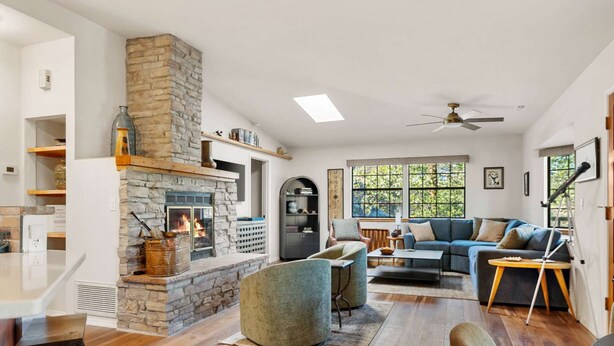 A living room with a stone fireplace, a ceiling fan, and a large window with a view of trees. - Sagewood Cottage: A private home with comfort, style, amazing amenities, and Tranquil location! (Prescott)