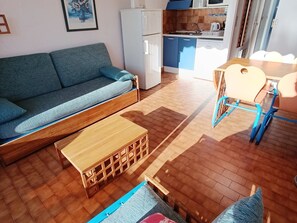 A compact living space with a sofa, a wooden table, a chair, and a kitchenette in the background. - Studio for 4 pers. with Wi-Fi, sea view (Roquebrune-Cap-Martin)