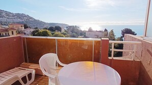 Un balcon avec une table ronde et deux chaises en plastique blanc, d’où l’on peut admirer une vue sur la côte, avec des bâtiments et des collines à l’horizon.