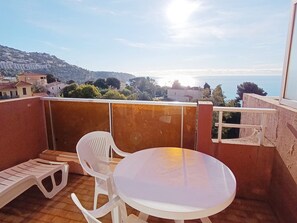 A balcony with a round table and two white plastic chairs, overlooking a coastal view with buildings and hills in the distance. - Studio for 4 pers. with Wi-Fi, sea view (Roquebrune-Cap-Martin)