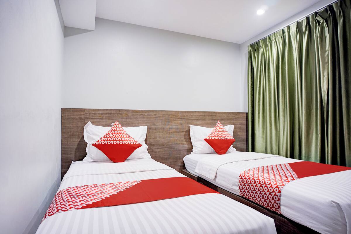 Habitación de hotel con dos camas, cada una con ropa de cama blanca y detalles en rojo, cabeceros de madera y cortina verde.