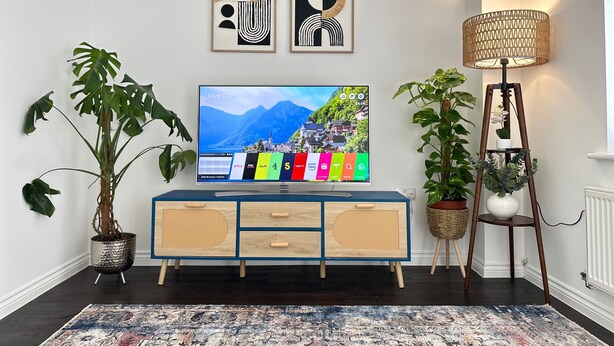 Una sala de estar moderna con un televisor mostrando una transmisión en vivo, un mueble azul de madera, dos obras de arte abstractas enmarcadas y plantas en macetas.