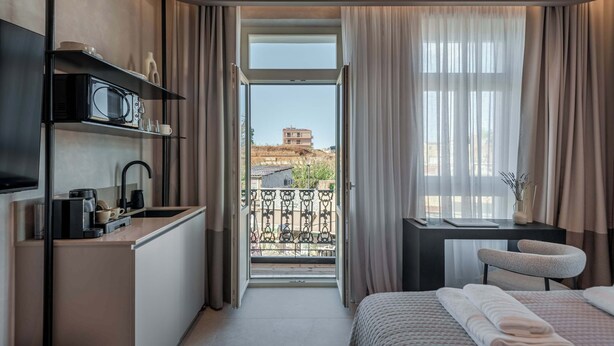 A compact hotel room with a kitchenette, a bed with neatly folded towels, a small desk with a chair, and a balcony with a view of a building.