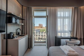 Habitación de hotel compacta con kitchenette, una cama y toallas ordenadas, un escritorio pequeño con silla, y un balcón con vista a un edificio.