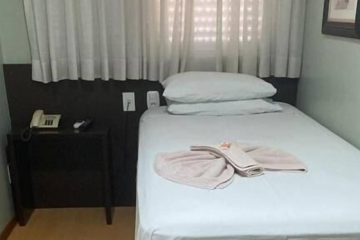 Ein Einzelbett mit weißer Bettdecke, ein Nachttisch mit Telefon und ein Fenster mit weißen Vorhängen.