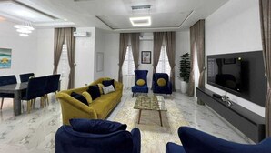 A modern living room with a yellow sofa, blue armchairs, a glass coffee table, a flat-screen TV, and a dining area with a table and chairs. - Welead Homes  (Abuja)