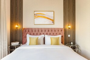 Een keurig opgemaakt bed met een geborduurde hoofdbord, twee nachtkastjes met lampen, en een lijst met een abstract schilderij aan de muur.
