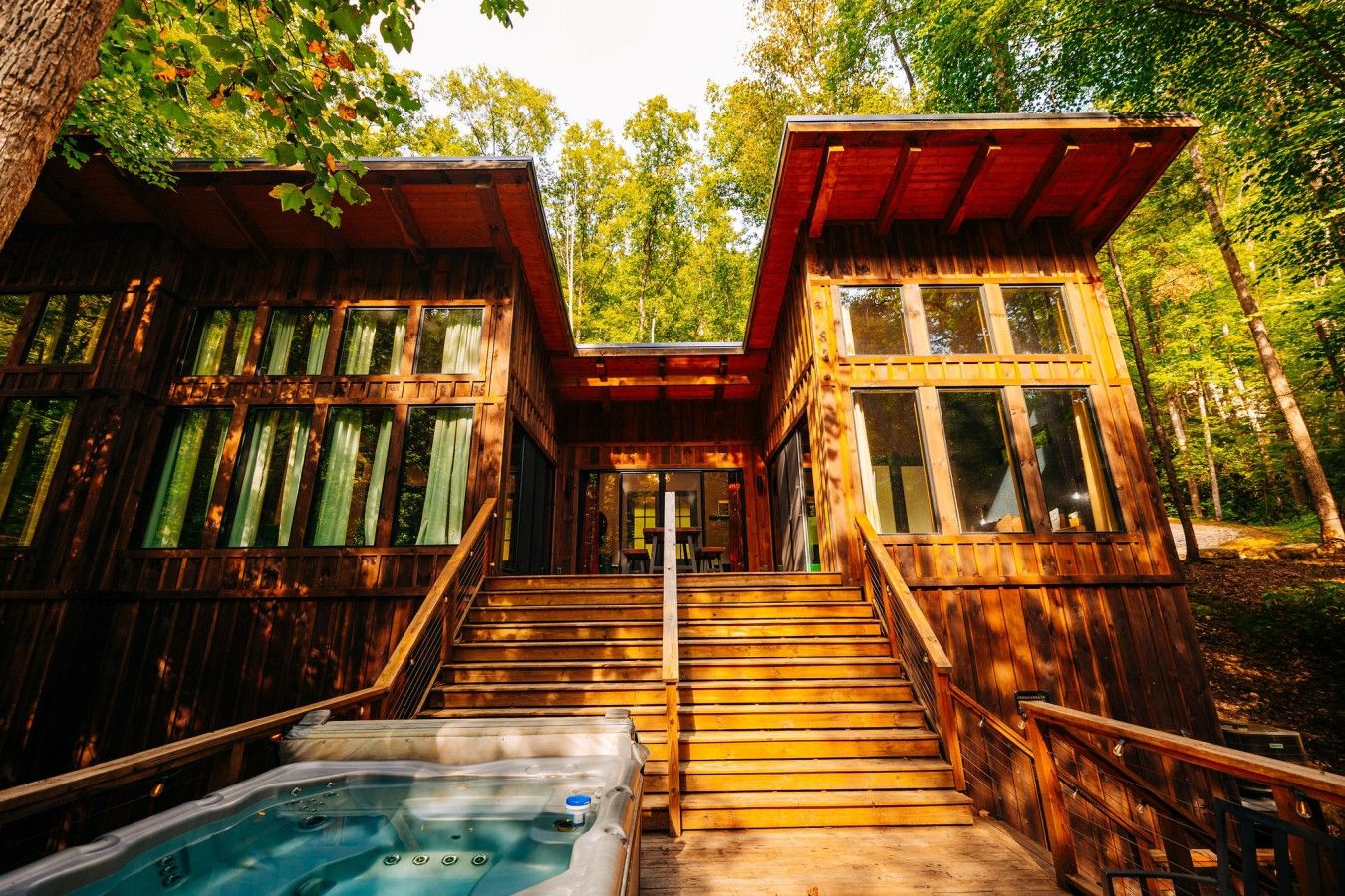 A wooden cabin with a hot tub, stairs, and a forested backdrop.