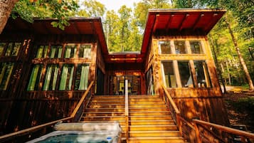 A wooden cabin with a hot tub, stairs, and a forested backdrop.