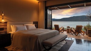 A bedroom with a large bed, a bench, two chairs, a nightstand, and a view of a lake and mountains. - Dali Kongshanyu Fengmian Seaview Hotel (Dali)