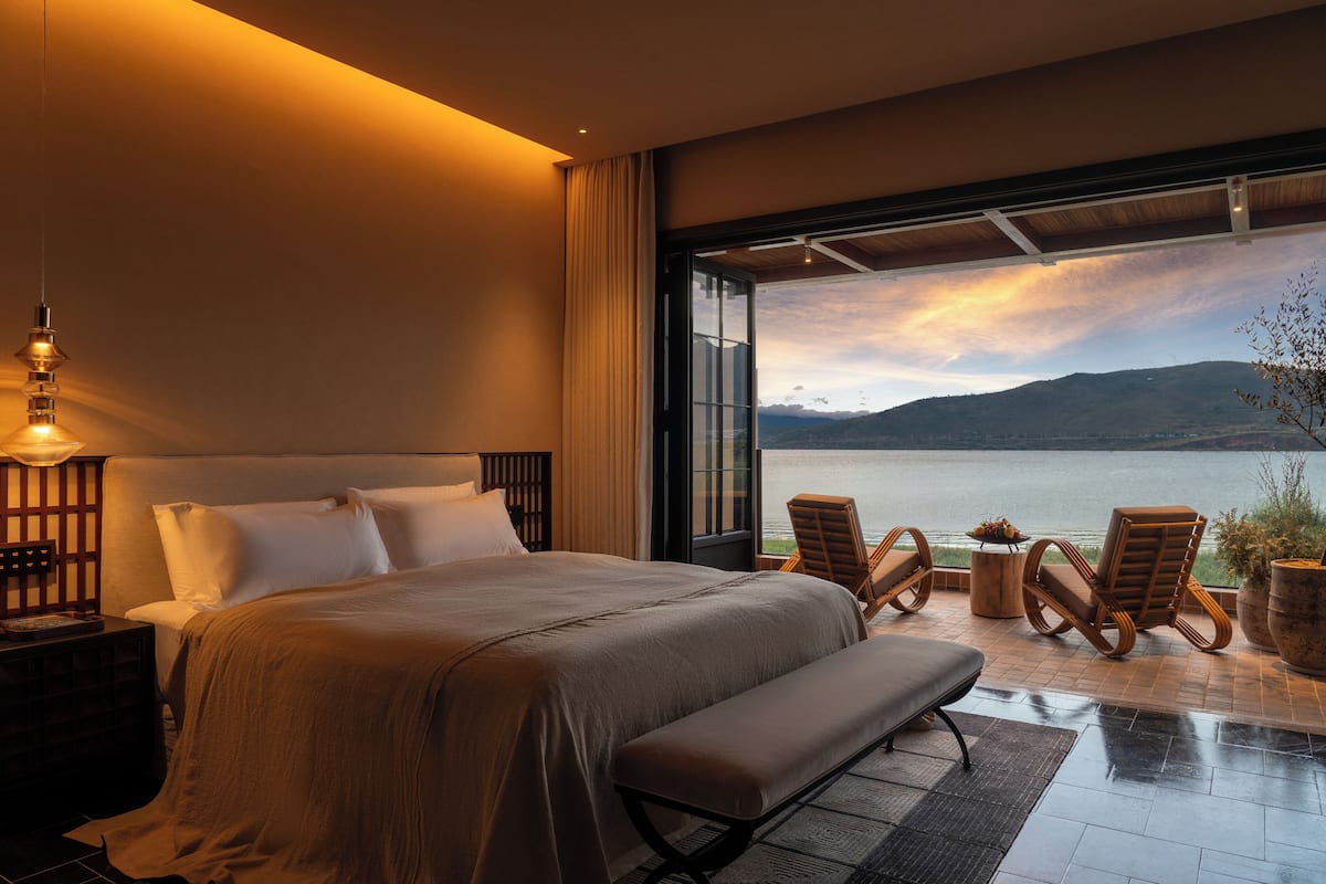 Un dormitorio con una cama grande, un banco, dos sillas, una mesita de noche y vistas a un lago y montañas.