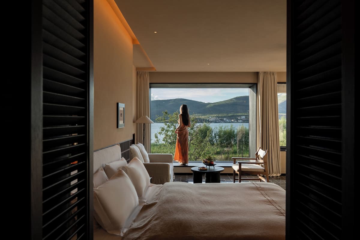 Habitación de hotel con una cama grande, zona de estar con una mesa y vistas a un lago y montañas.