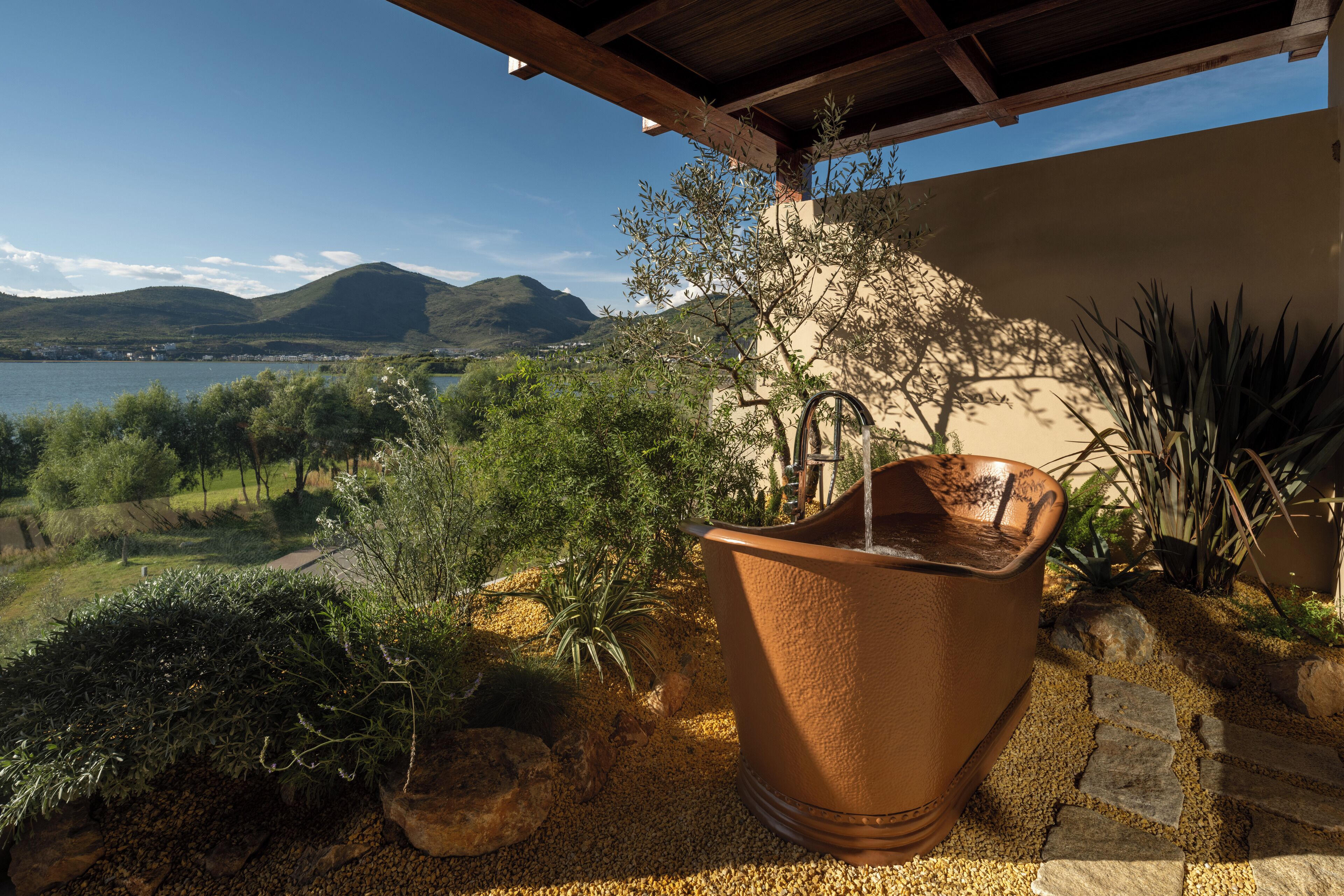A terracotta pot with water, surrounded by rocks and plants, overlooking a scenic landscape with hills and a body of water.