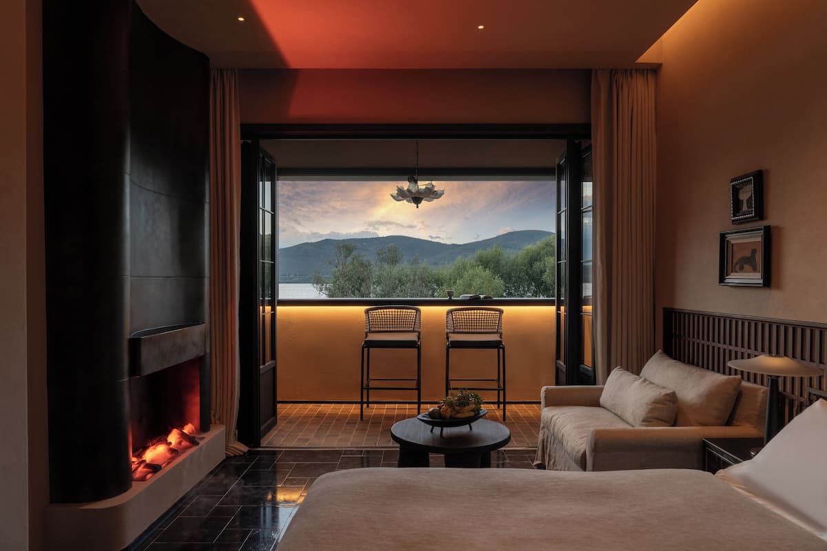 Una habitación de hotel moderna con chimenea, un ventanal con vistas a la montaña, dos sillones, un sofá y una mesita.
