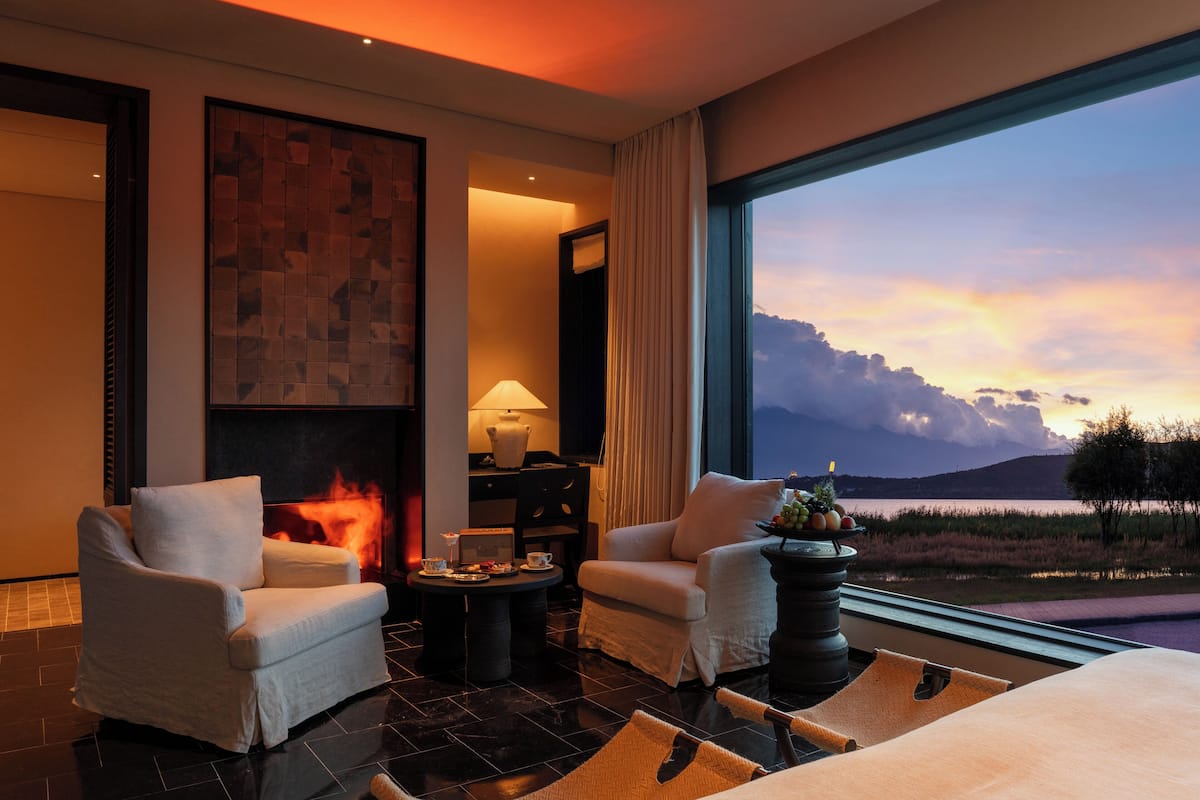 Habitación de hotel moderna con chimenea, dos sillones, una mesa de centro y un amplio ventanal con vistas panorámicas del paisaje.