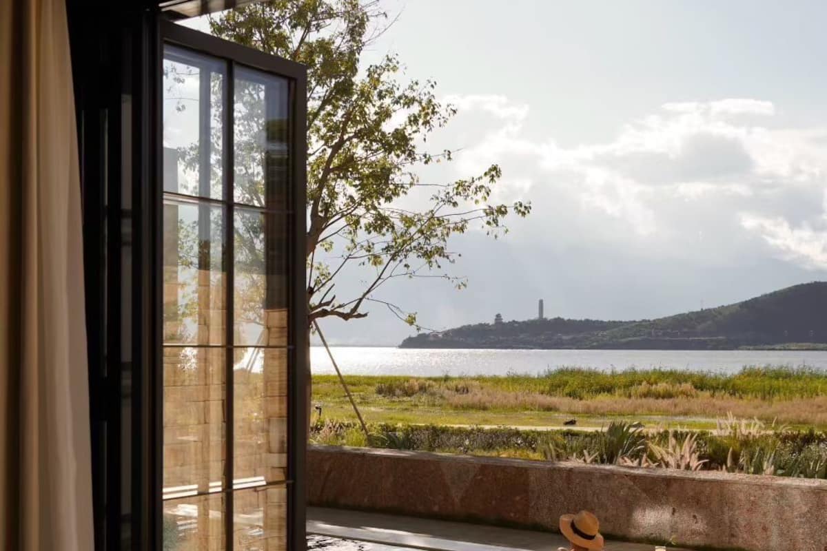 Una habitación con un amplio ventanal que da a una piscina, vista de una colina distante y una mesa con frutas y una bebida.