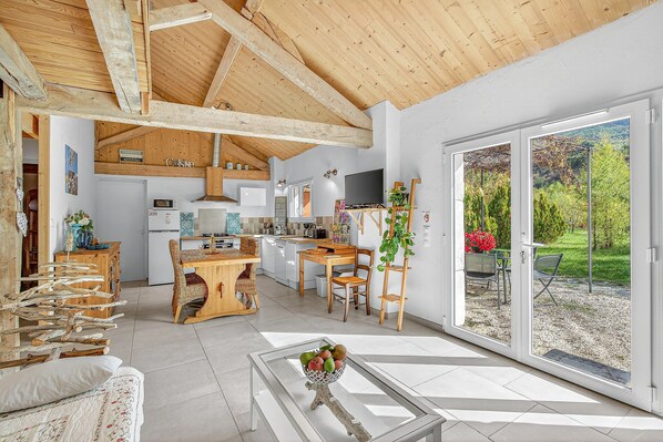 A spacious living area with a high ceiling, wooden beams, a glass coffee table, and a sliding glass door leading to a garden.