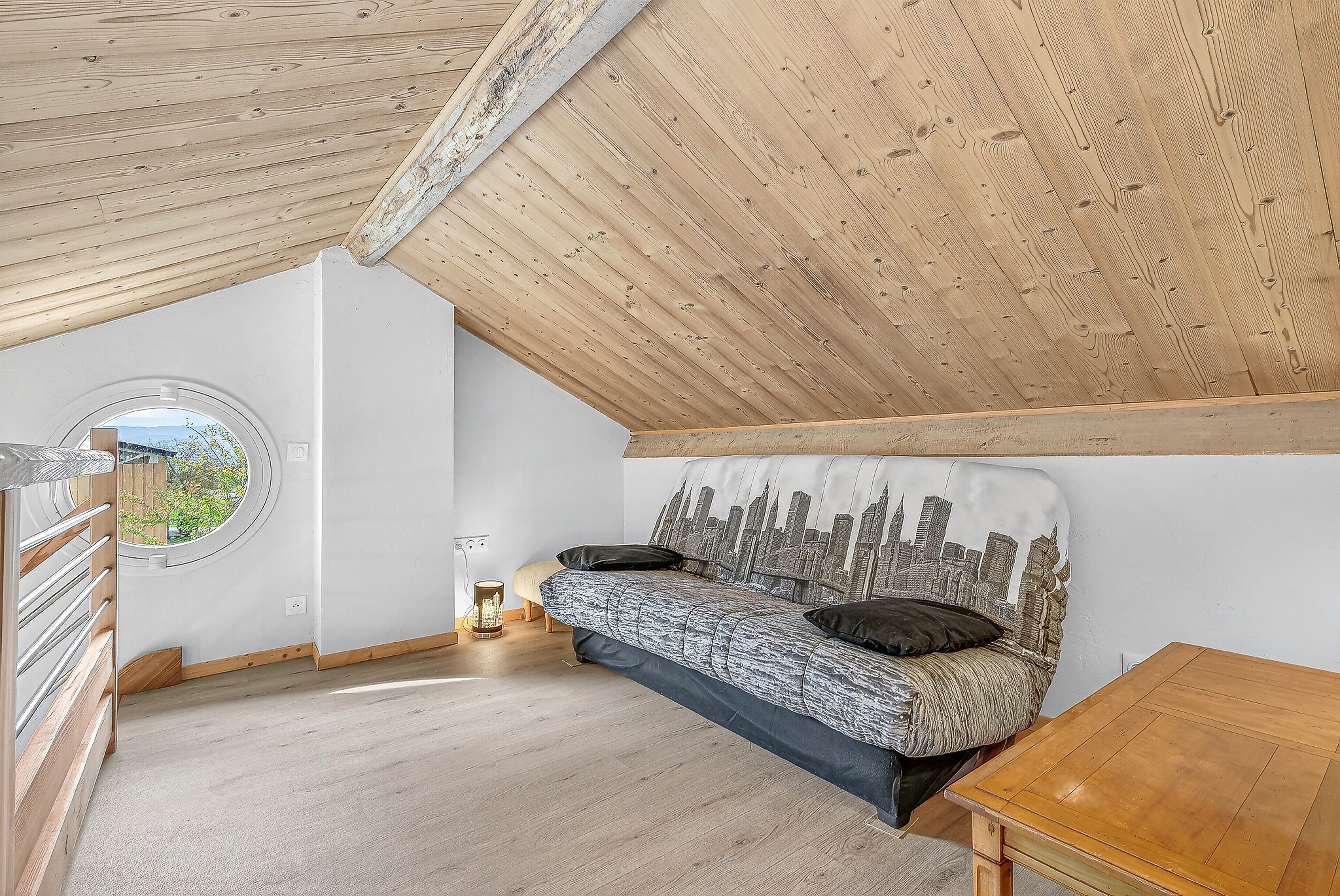 A loft bedroom with a sloped ceiling, a cityscape headboard, a circular window, and a wooden floor.