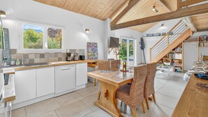 Une cuisine spacieuse avec des meubles blancs, une table à manger en bois et une vue sur l’extérieur grâce à de grandes fenêtres.