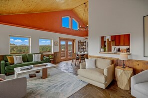 A spacious living area with a high ceiling, a green sofa, a beige armchair, and a dining area with wooden furniture. - Retro-Modern, Mountain Retreat with 3 bedrooms, 2 baths, a bonus game room, office, and a beautiful deck with ample seating for enjoying the Stunning Mountain Peak Views. Best Views in town. (Pagosa Springs)