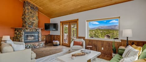A cozy living room with a stone fireplace, comfortable seating, a flat-screen TV, and a large window offering a scenic view.