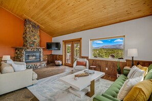 A cozy living room with a stone fireplace, comfortable seating, a flat-screen TV, and a large window offering a scenic view.