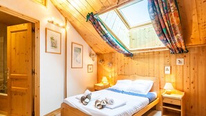 A wooden room with a sloped ceiling, a bed with white linens, a nightstand with a lamp, and a skylight with curtains. - Spacious 3-bedroom ski-in apartment with parking (Méribel)