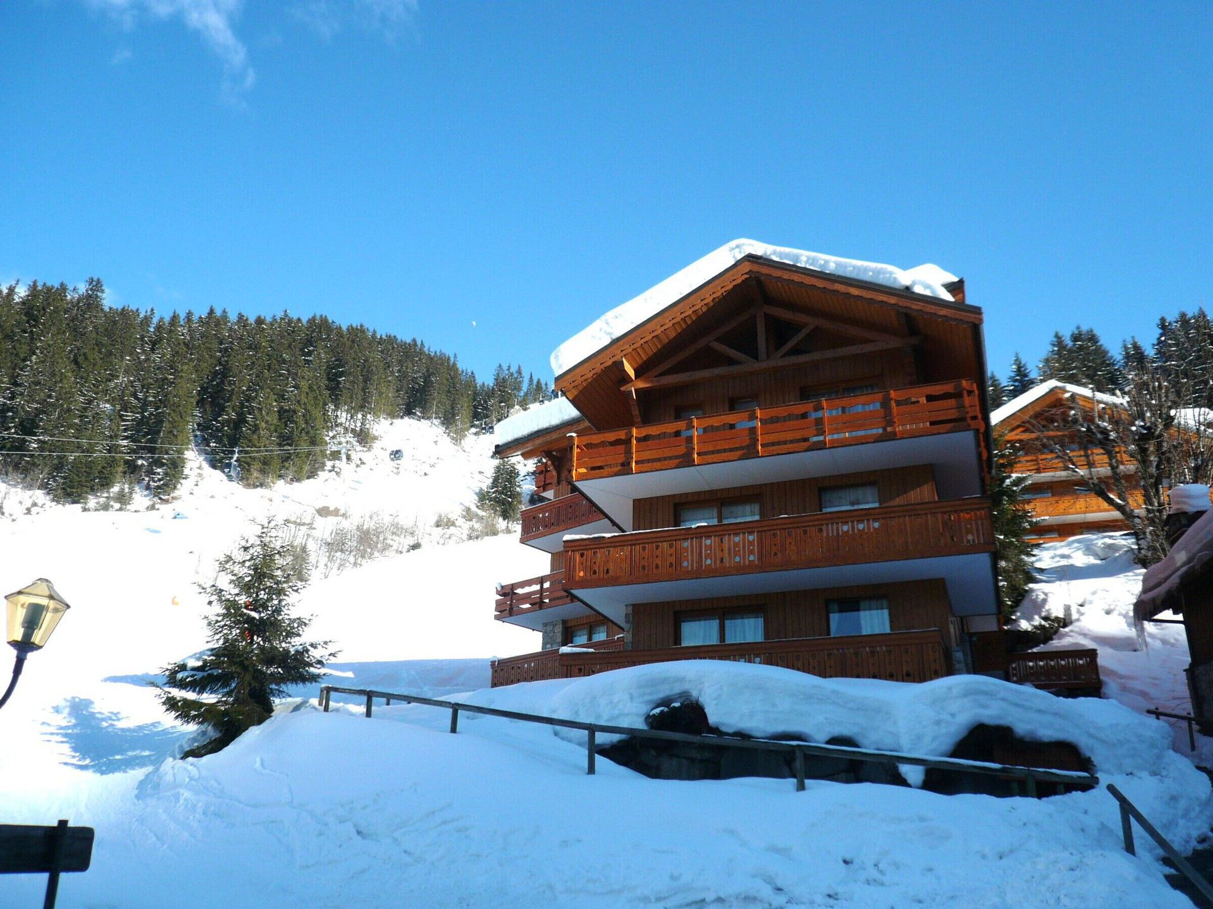 A multi-story wooden chalet with balconies, surrounded by snow-covered trees and a clear blue sky.
