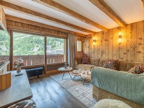 A cozy room with a wooden ceiling, a sofa with patterned cushions, a small round table with cups, and a view of a forested area through large windows.