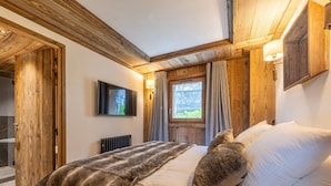 A bedroom with a wooden ceiling, a bed with a fur throw, a stone wall, and a window with curtains.