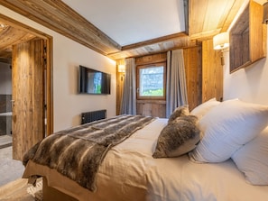 A bedroom with a wooden ceiling, a bed with a fur throw, a stone wall, and a window with curtains.