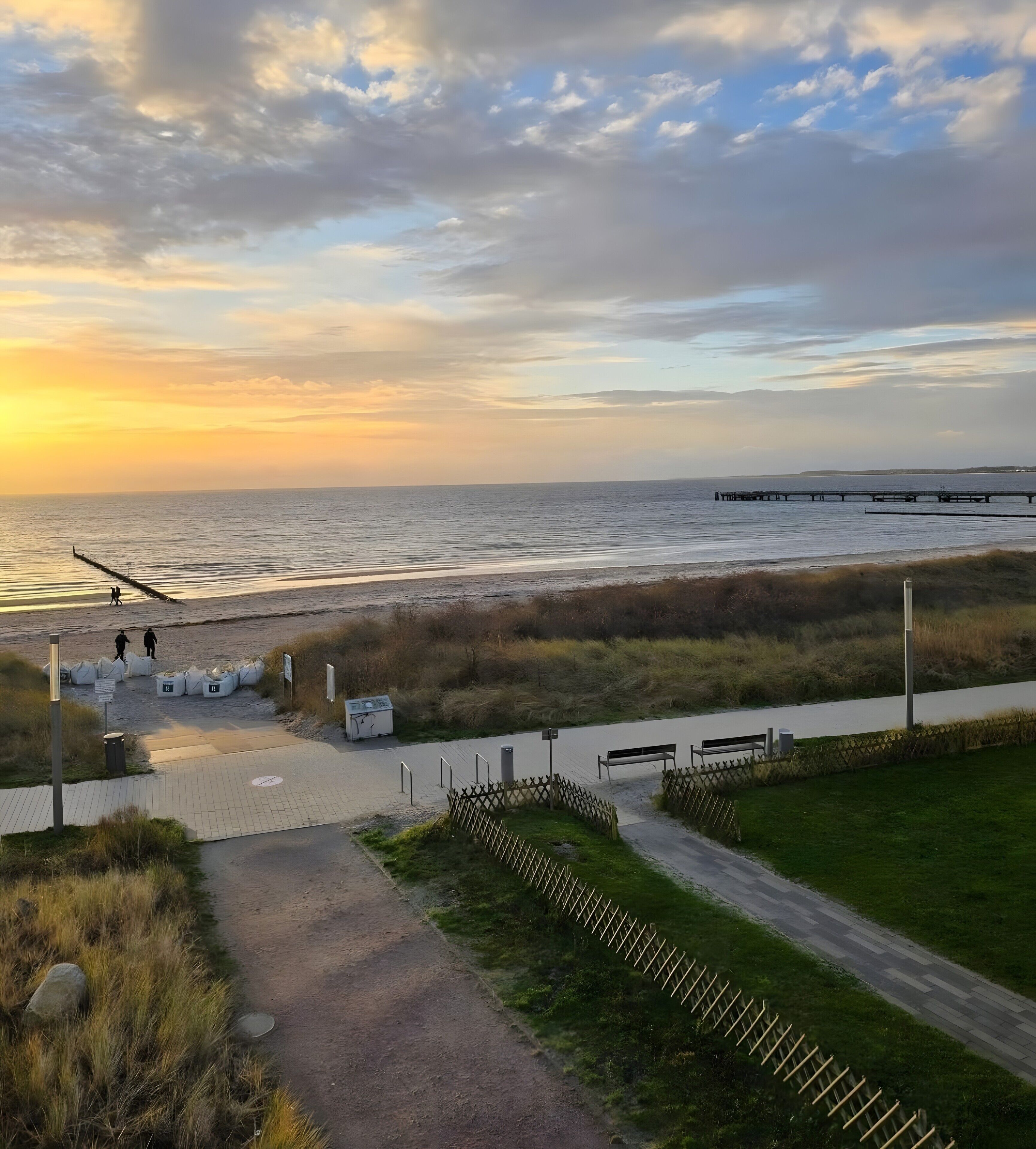 Ein Küstenweg mit Blick auf das Meer, ein Pier in der Ferne und eine Rasenfläche mit Bänken.