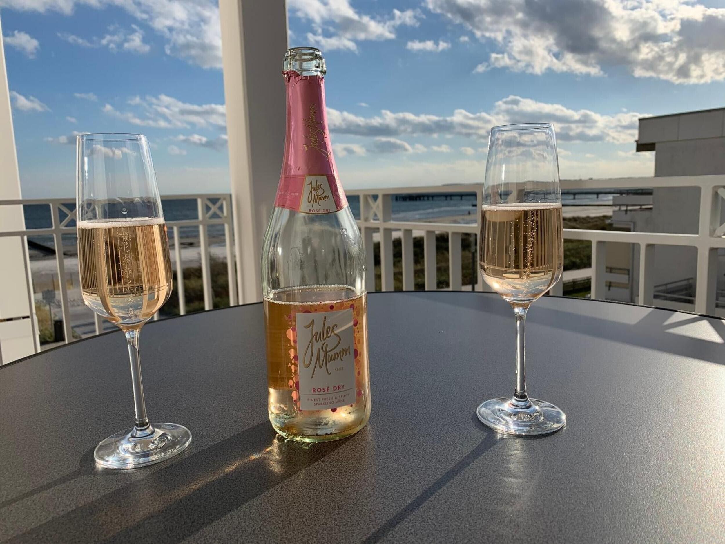Jules Mumm Rose Dry -kuohuviinipullo ja kaksi lasia pöydällä, josta avautuu näkymä merelle.