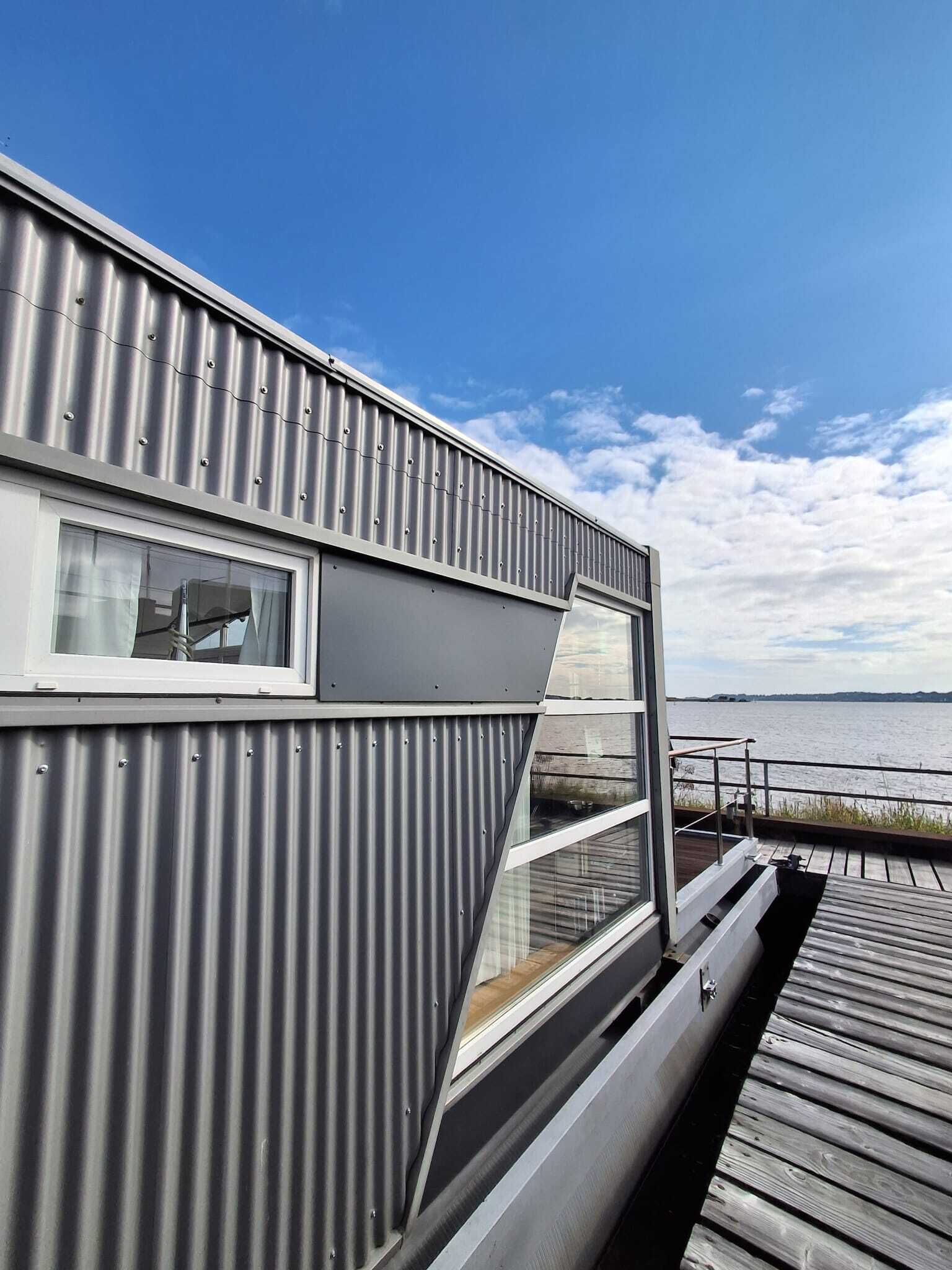 A modern building with a corrugated metal exterior, a large window, and a wooden deck overlooking a body of water.