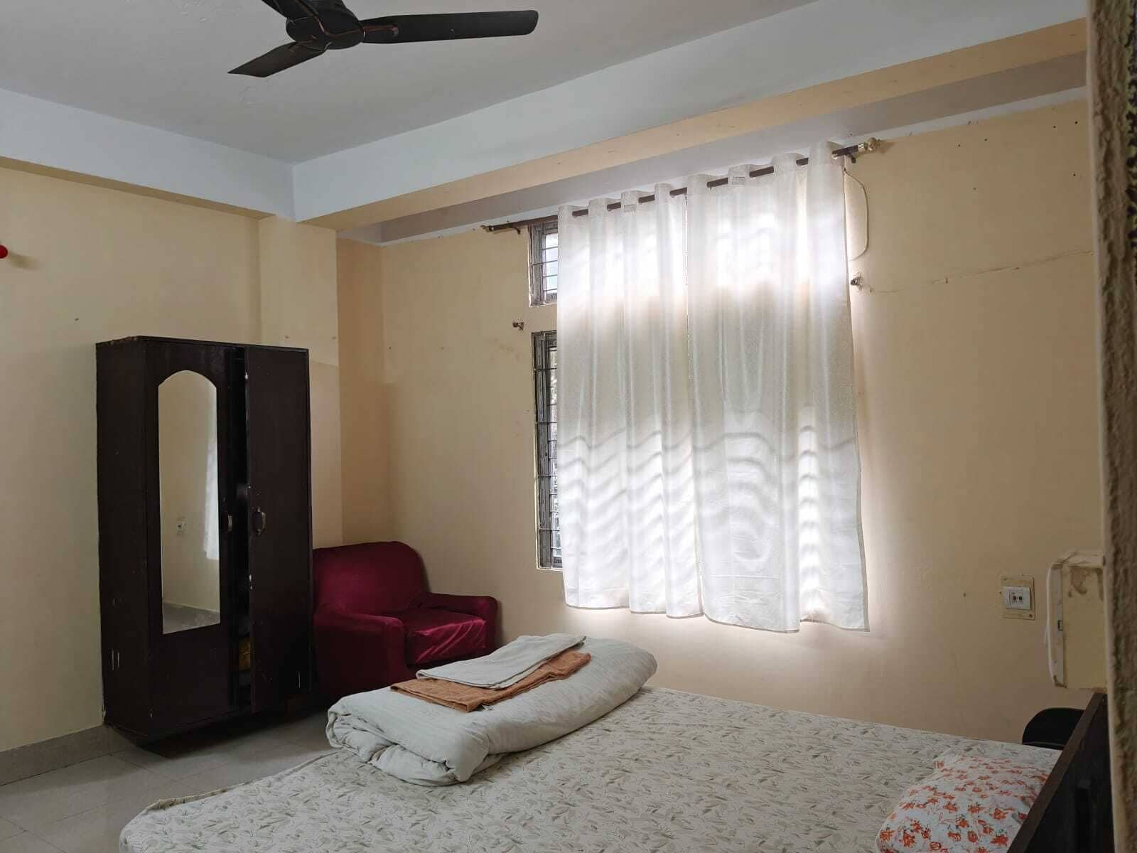 Une chambre à coucher avec un lit, une armoire, une chaise rouge, une fenêtre avec des rideaux et un ventilateur de plafond.