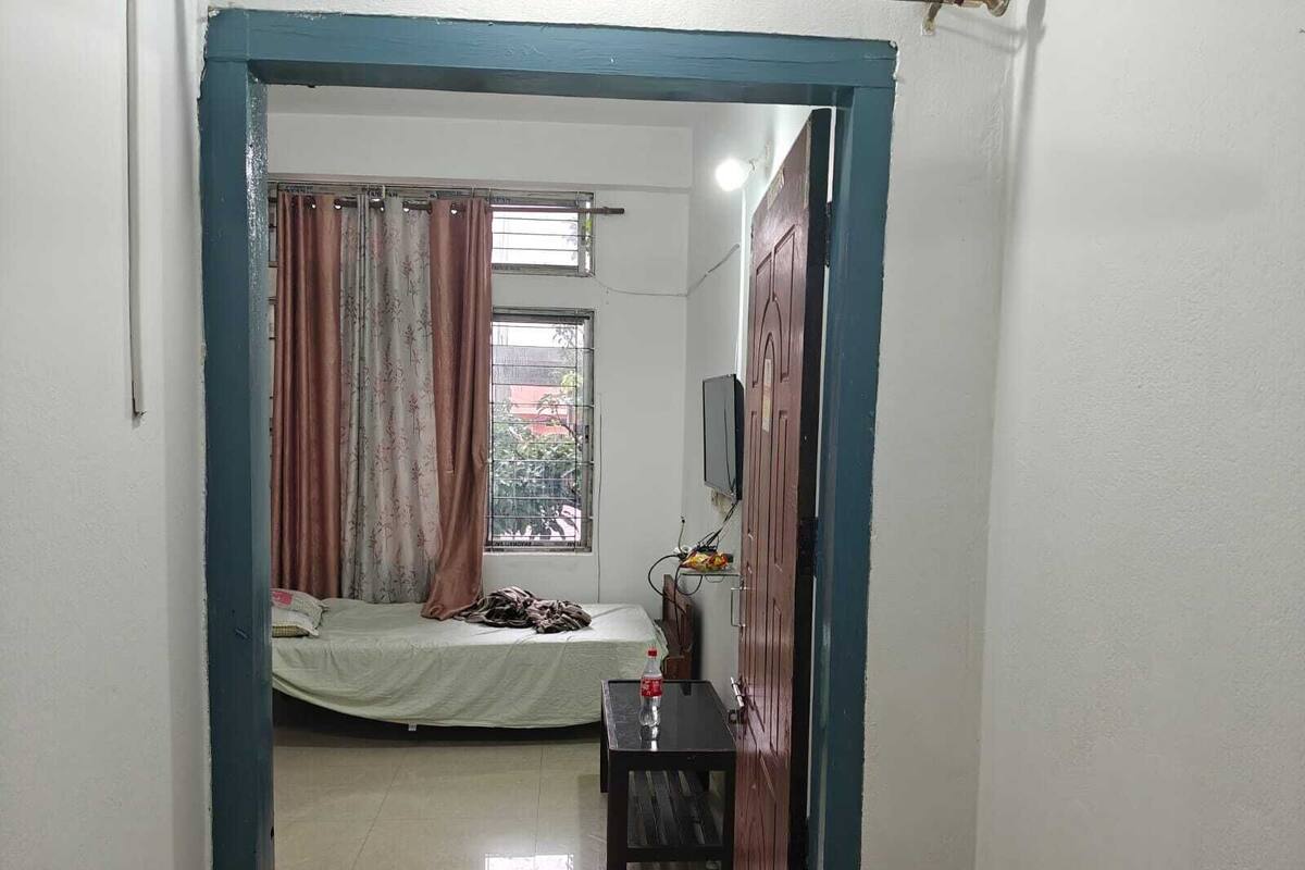 A small, well-lit room with a bed, a TV mounted on the wall, and a window with curtains.