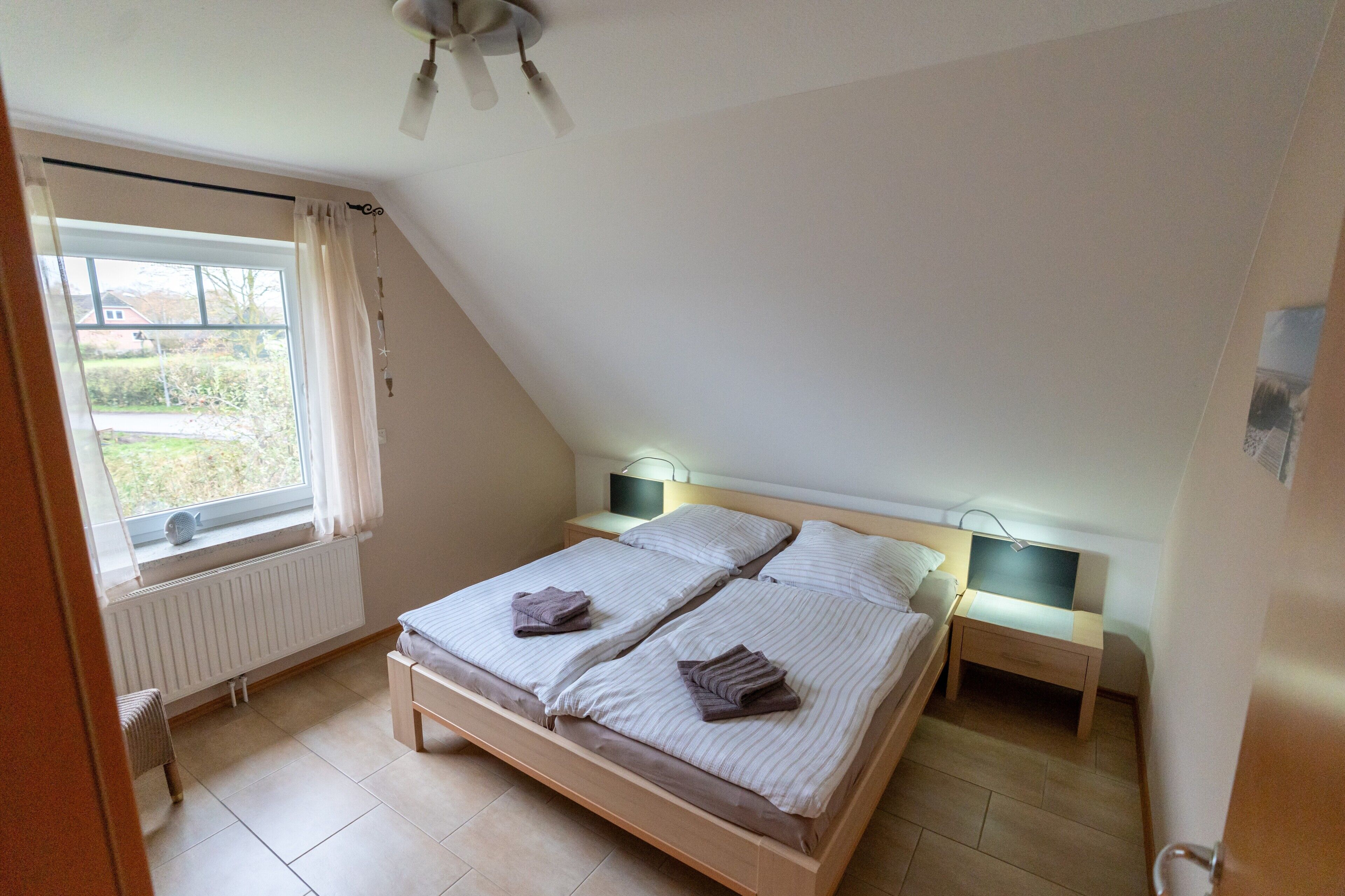Ein Schlafzimmer mit schrägem Dach, zwei Betten, Nachttischen, einem Heizkörper und einem Fenster mit Vorhängen.
