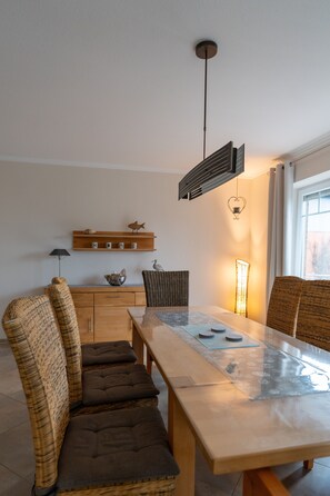 A dining area with a wooden table, wicker chairs, a hanging light fixture, and a shelf with decorative items.