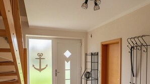 Un pasillo con escalera de madera, una puerta decorada con un ancla, un perchero metálico y una lámpara en el techo.