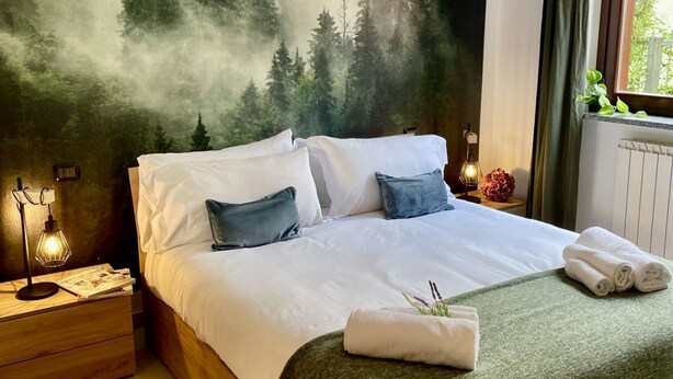 A neatly made bed with white and blue pillows, a green throw blanket, and rolled towels on a wooden nightstand. A wall-mounted lamp and a small table with a candle are visible.