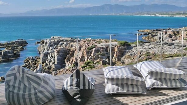 A deck with striped cushions and a view of the sea and rocky coastline. - Seafront Villa - Whale Bay Luxury Retreat (De Kelders)