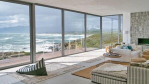 A modern living room with large floor-to-ceiling windows, a white sofa, a coffee table, and a stone wall. - Seafront Villa - Whale Bay Luxury Retreat (De Kelders)