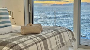 A bedroom with a large window overlooking the sea, a bed with a plaid blanket, and a bedside lamp. - Seafront Villa - Whale Bay Luxury Retreat (De Kelders)