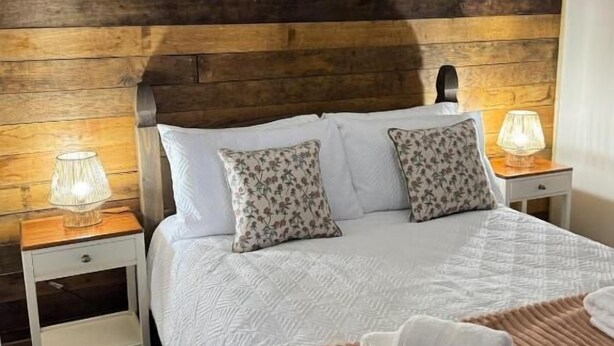 A bedroom with a wooden headboard, white bedding, two floral pillows, a bedside lamp, and a folded towel on the bed. - The Riley Stable- Cozy Getaway (Waterlooville)