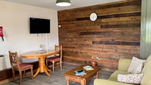 A cozy living room with a wooden wall, a round wooden table with chairs, a television mounted on the wall, and a green hanging light fixture. - The Riley Stable- Cozy Getaway (Waterlooville)